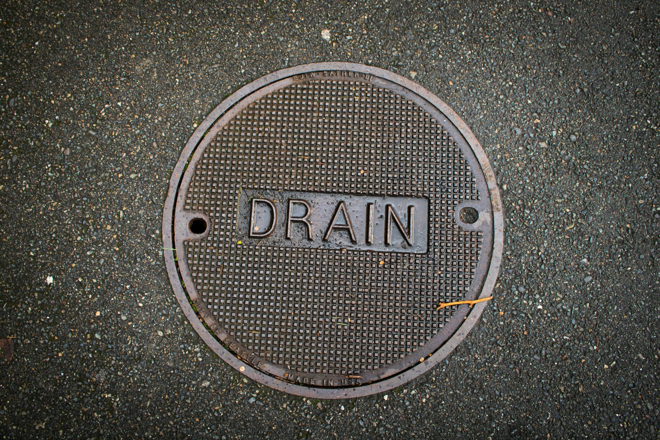 This close-up photograph shows a round metal manhole cover, clearly labeled with the word "DRAIN," set within an asphalt surface. The cover features a textured grid pattern, providing visual interest and likely enhancing grip.