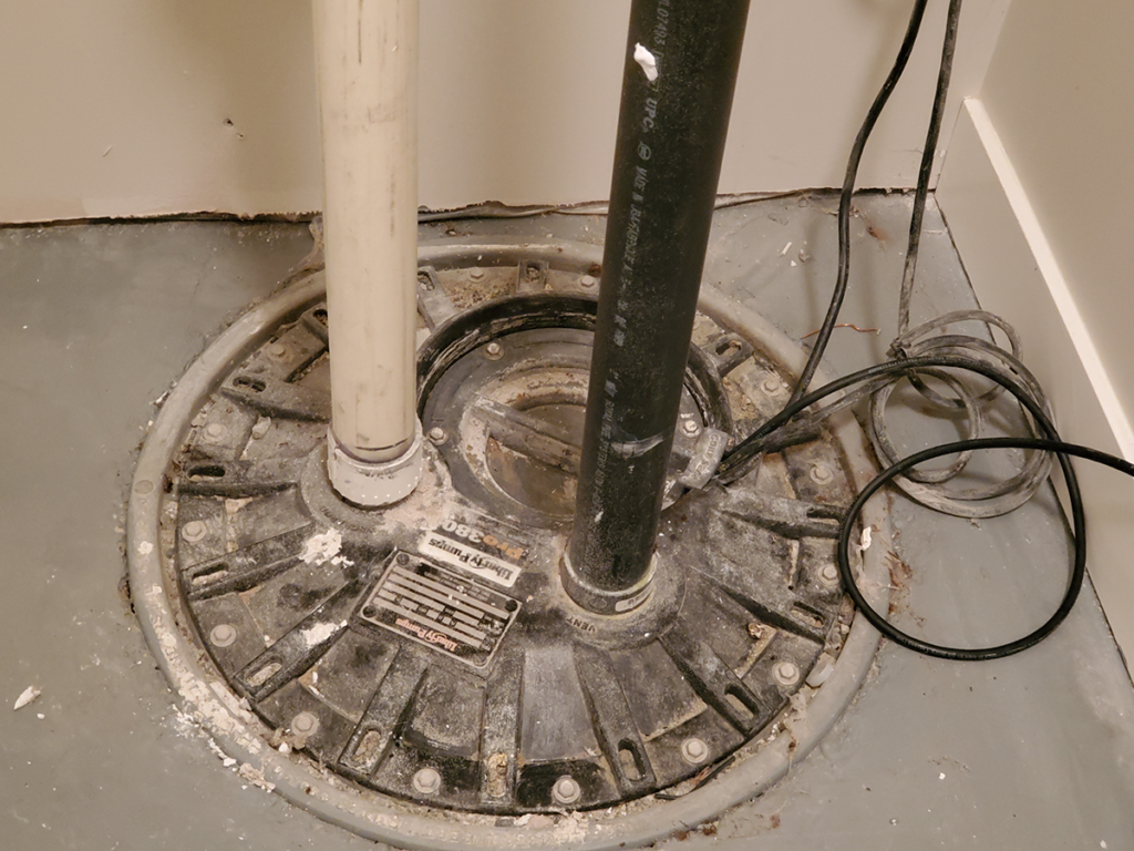 "Image of an installed recessed and embedded sump with two vertical pipes and electrical wiring, set in a concrete floor."