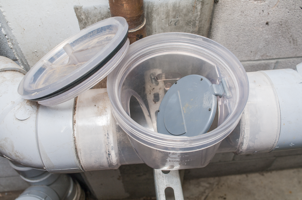 Image of a backwater valve installed in a plumbing system, with the transparent cover removed to show the internal flap mechanism that prevents backflow in the pipes.
