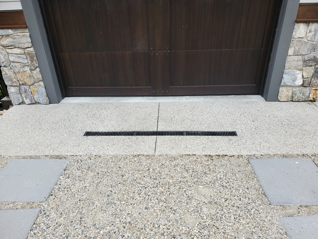 "Image of a trough or trench drain installed in front of a garage, embedded in the concrete driveway."