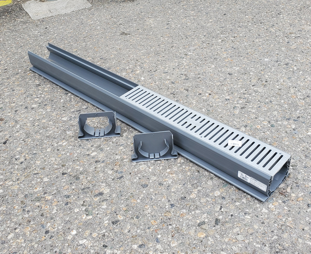 "Image of a plastic trough drain with a grated cover, placed on a concrete surface, alongside two end caps."